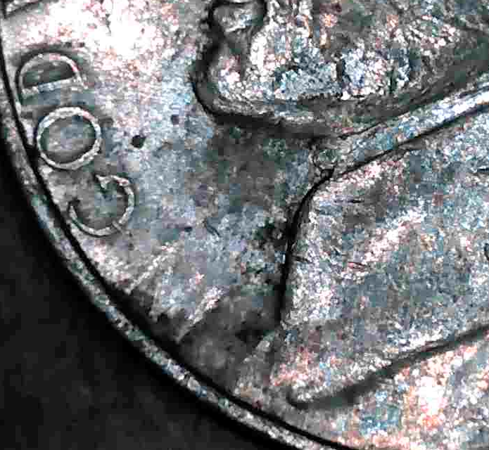 A close-up of a 1999 Jefferson nickel showing the words IN GOD partially missing due to a grease-filled die, with smeared detail on Jefferson’s jaw and collar.