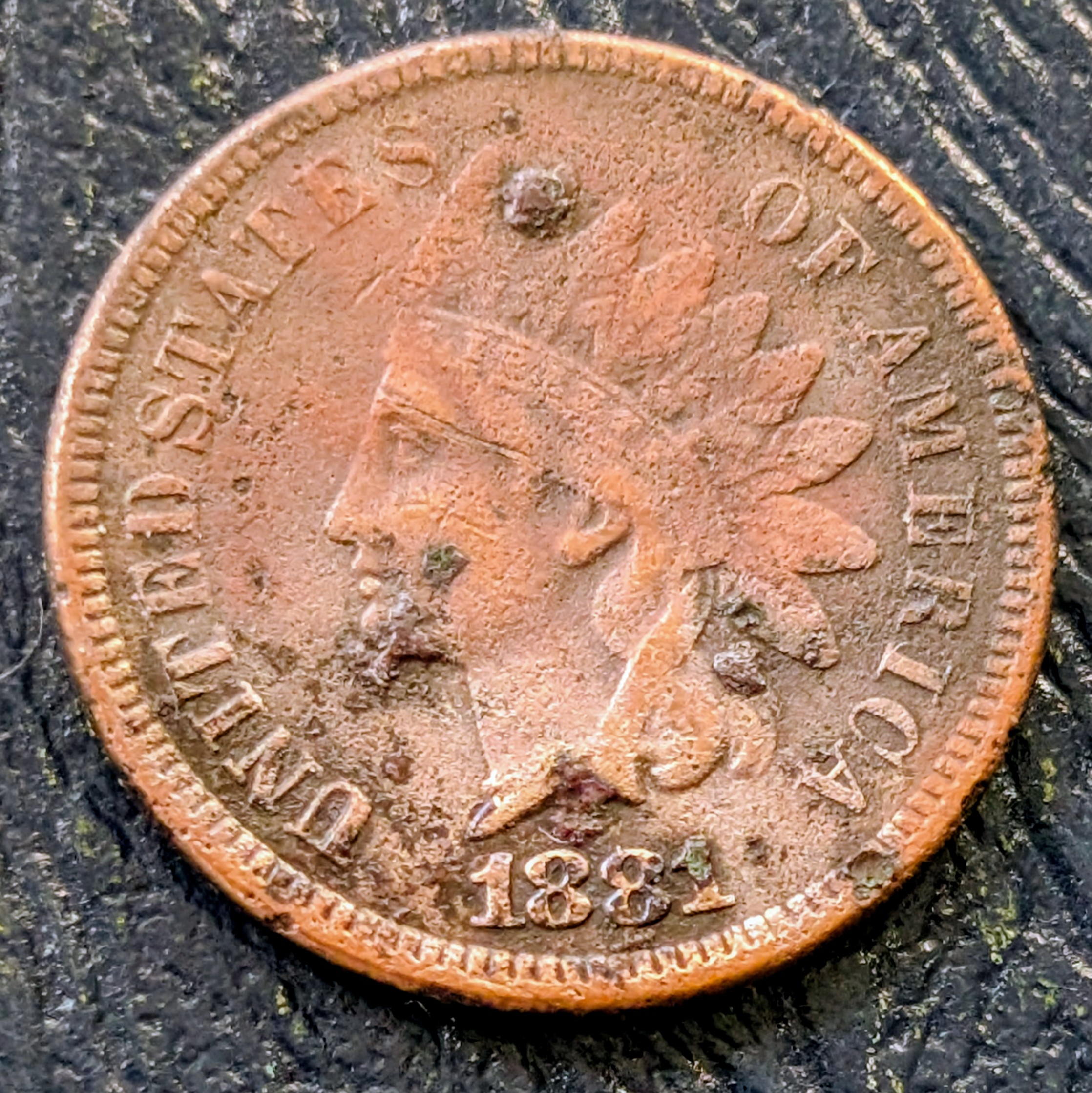1881 Indian Head Cent with heavy environmental damage showing green and brown corrosion, missing surface metal, rough texture, and uneven crust across the portrait and fields.