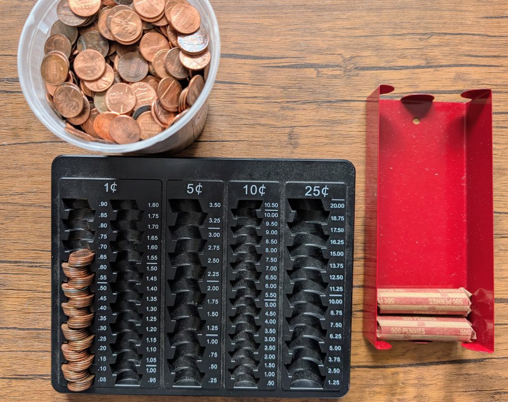 A tabletop coin rolling setup with a container of loose pennies, a black plastic coin counting tray, and a red metal holder with paper coin rolls.